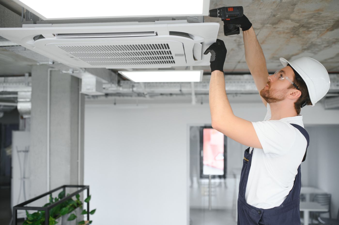 Pracownik budowlany w białym kasku i kombinezonie instaluje lub naprawia jednostkę klimatyzacyjną sufitową za pomocą wiertarki. Sceneria wydaje się być niedokończoną przestrzenią wewnętrzną z widocznymi kanałami i elementami betonowymi.