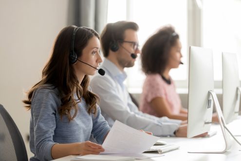 Trzech pracowników call center w słuchawkach siedzi przy biurkach przed komputerami. Na pierwszym planie widać kobietę trzymającą papiery, podczas gdy dwaj koledzy w tle skupiają się na swoich ekranach. Jasne światło dzienne sączy się przez okno, a powietrze jest chłodne i świeże dzięki wydajnej klimatyzacji.